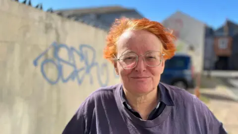 George Carden/BBC Rosemary who has orange dyed hair standing in front of a wall near her house with blue spray paint on it