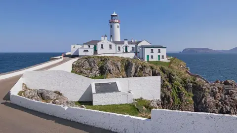 Getty A large white building with a number of doors and windows sits on top of a cliff face. A red and white lighthouse is visible over the structure.
A large body of water and rocks are visible in the background
