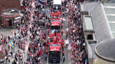 Fans in red and white shirts swarm around open top buses
