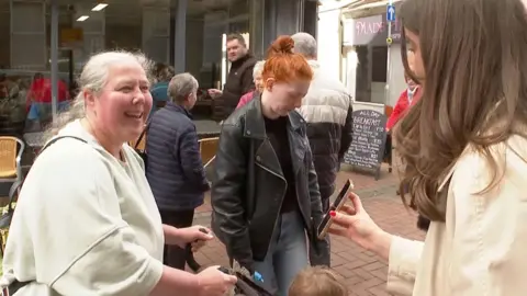 A woman with tied-back grey hair, a light green sweatshirt and a black backpack is pushing a pram and smiling as she looks at a reporter whi has long brown hair and is holding out her phone to the woman