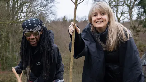 The Woodland Trust Nile Rodgers and Nancy Hunt plant a tree in woodlands near Derry. Nancy, with blonde hair and wearing a black coat is holding a spaling. Rodgers is on the left and wearing dark glasses , a head scarf and cblack jacket, and is holding a spade.