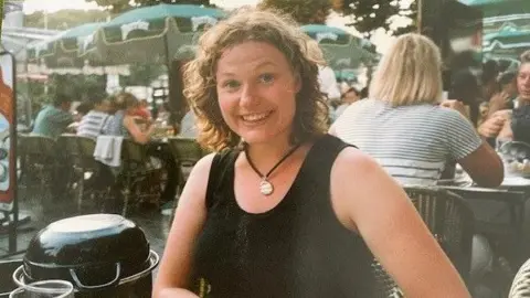 A woman sits at a table in a Parisian outdoor cafe and smiles for the camera. She is suurounded by people seated at other tables. She has curly brown hear and wears a black sleeveless top and a gold pendant around her neck.