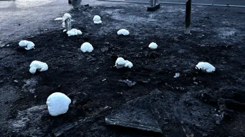 George King/BBC The minimal remains of the damaged play area. Very little remains and the pieces of wood that do are covered with some type of white, protective foam. The area has been noticeably heavily charred by the fire. 