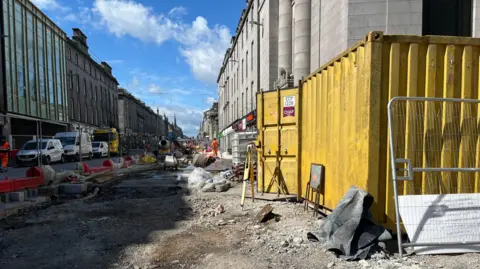 BBC Union Street on a bright but cloudy day. Old granite buildings are to the side of the road, which has been dug up. There's a yellow container to the left of the shot. 