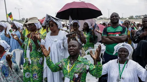 AFP via Getty Images A man in a vibrant green shirt with his eyes closed and hands raised in prayer. He is kneeling as is the woman on his left. Behind them are dozens of people standing, including one with a cap on, one with an umbrella up, and a woman with an open book over her head, all to shield from the sun.