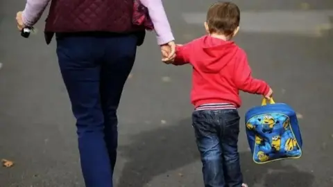 The back of a woman holding a young boy's hand. She is wearing blue jeans and holding car keys in her other hand. The boy is wearing a red hooded top and blue jeans, and is holding a yellow and blue minions backpack.