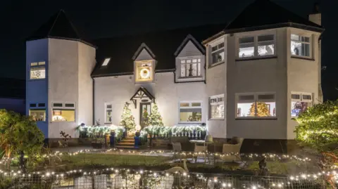 Kirsty Anderson White two-story house at night with Christmas lights, wreath, and decorated trees in front garden. There is a pond with fairy lights around it.