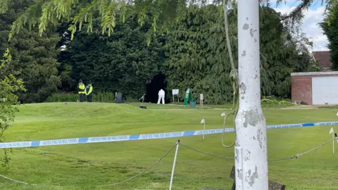 Police tape at the golf course behind a tree. Figures can be seen in the distance around a path.