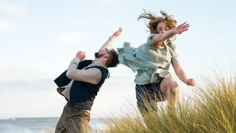 Charlie Dunne and Lily Horgan are dancing on the coast. Charlie has his hand raised and is back arched. Lily is mid jump and is reaching towards the camera. The ocean is in the background and grassy dunes are in the foreground.