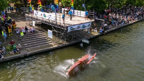Home made plane contraption falling into water from a low platform. Lots of people are on the banks of the harbour watching.
