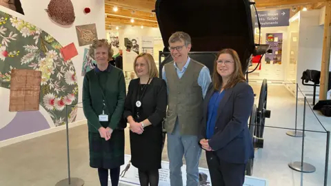 Three women and a man stand in front of a museum display while smiling for the camera. Artwork can be seen on the wall to their right. 