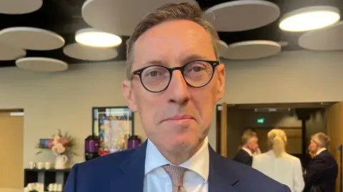 BBC Ian Gorst has short dark hair, is wearing glasses, and a blue jacket with a white shirt and light coloured.