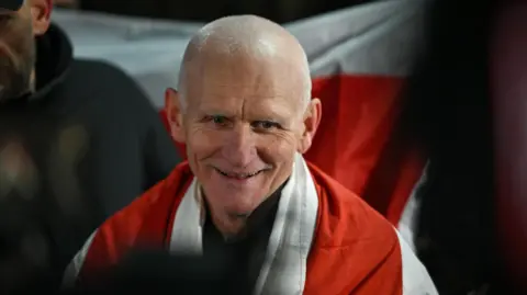 EPA/Shutterstock Belarusian social activist, politician, human rights defender and political prisoner Ales Bialiatski in front of the US Embassy in Vilnius, Lithuania. He is a man in his 60s, smiling and has close-cropped white hair. Around his shoulders is the red and white opposition flag of Belarus.