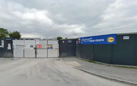 Google Entrance to a construction site secured by a metal gate and dark hoarding panels under a cloudy sky. Safety signs are fixed to the gate, and a large blue Lidl banner on the right reads “We’re cracking on with your new store.” The ground is a dusty access road leading up to the closed site.

