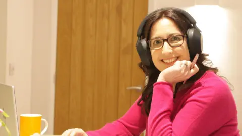 Contributed Christina Reeves sits at a table with headphones on and a laptop in front of her. She is looking to the left toward the camera and is smiling. She has one hand that holds a pen resting on her chin. She is wearing a bright pink long sleeved top.