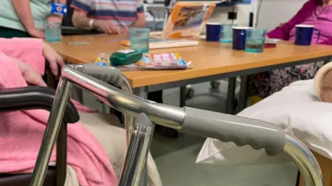 Getty Images A stroller in front of a table with elderly people in a care home setting. On the table cups with drinks, and magazines for them to read. Several old people are sitting around the table. 