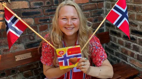 Ragnhild Lund Ansnes has long blonde hair and is smiling into the camera. She is wearing a red floral dress while carrying a packet of Norwegian chocolate and two Norway flags.