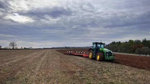 A green tractor is ploughing a field. On the left is the untouched grass, on the right you can see the soil that has been turned over. The sky is a little foreboding and full of clouds.