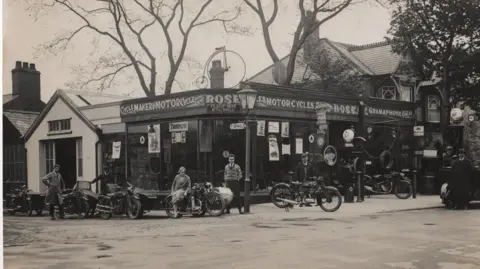 March and District Museum A black and white shot of the front of the garage in 1930. There are motorcycles on the forecourt with a petrol pump on the right. A woman is sitting side-saddle on a motorbike and some men are standing around. A Penny Farthing sits on top of the shop.