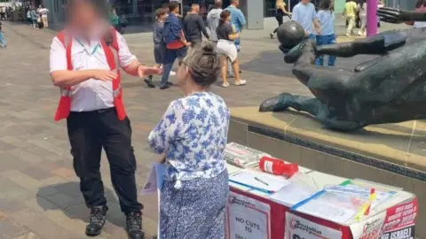 A city warden in a red and black uniform talks to a woman in a flowery blue top who is standing by a table in the street