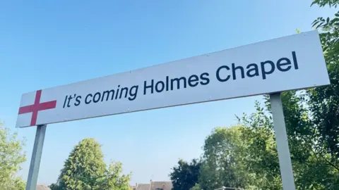 Northern Railway station sign reading: 'It's coming Holmes Chapel'.