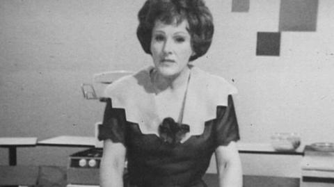 Black and white picture of Fanny Cradock on set in her kitchen studio. Wearing a black dress with white collar.  Kitchware and products visible in the background of the shot.