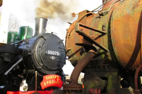 North Dorset Railway Two steam locomotives stand nose-to-onse. One is completely restored with new black and green paint and steam is coming out of its chimney, the other is covered in surface rust.