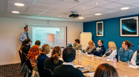 Office of the Police and Crime Commissioner for Cleveland A group of people are sitting round a table in a hotel conference room receiving training. A man in a shirt and tie is at the front standing in front of a projected Powerpoint slide.