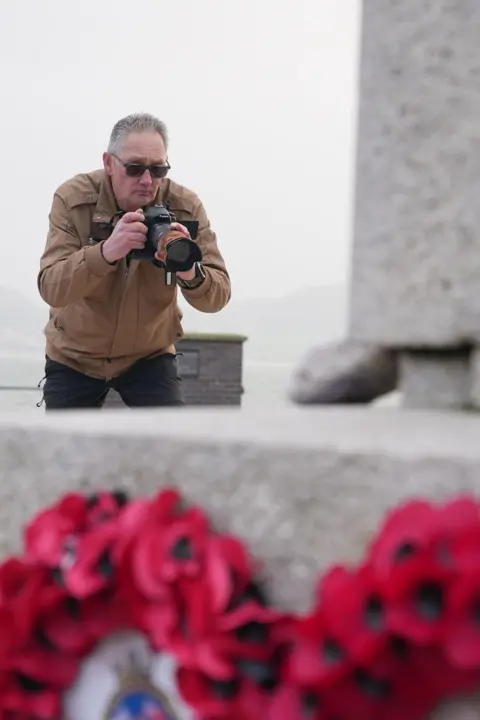 A white-haired man in a brown jacket wearing dark glasses is leaning forward with a camera taking a picture of a grey cenotaph with a wreath of poppies in the foreground