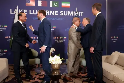 Reuters All four men, wearing suits, greet each other in front of an official sign for the summit. 