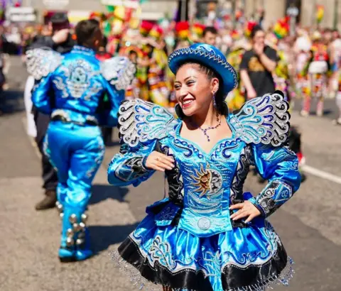 A parade participant in an ornate bright blue costume with silver embroidery and a matching hat performs on a city street during a colourful outdoor festival. Other costumed performers and spectators are visible in the background.