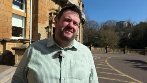 Rich Herrett is standing outside a limestone building. He has short black hair and is wearing a green and white striped shirt.