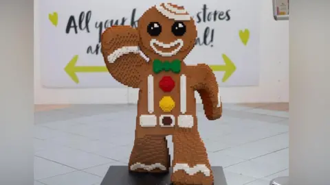 A large figure of a gingerbread man created out of Lego bricks. The colouring is brown with round black eyes, and a white outline of a mouth, and one arm raised. A green bow tie, and a red and yellow button complete the "outfit". It is standing in what appears to be an indoor shopping mall.