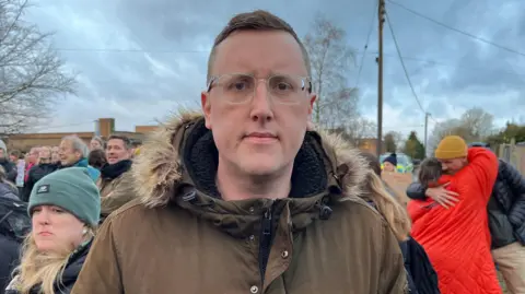 Shaun Whitmore/BBC Man with glasses wears a warm brown coat. Next to him is a woman with a blue hat. In the background are more adults. All are outside a school. 