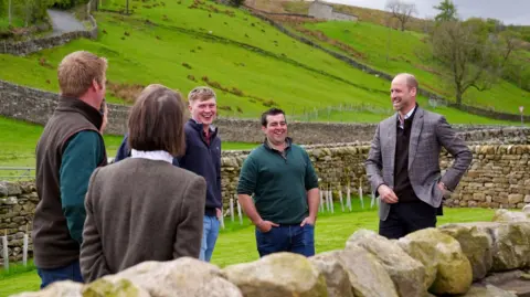 Owen Humphreys/PA The Prince of Wales stood with a group of five farmers. The scenery in the background is hills and lush green fields. He is wearing a grey suit jacket and has a hand on his hip. The farmers are mostly wearing blue jeans and brown or green quarter zip jumpers. They are all laughing.