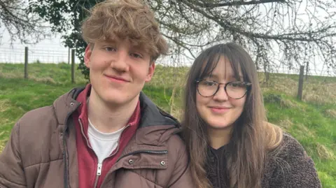A young man and woman smiling at the camera. He has blond hair and a brown padded jacket, while she has brown hair with a fringe, is wearing glasses and a fluffy brown coat.