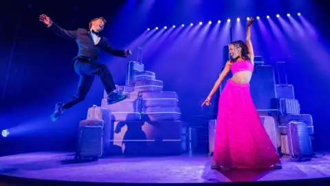 Matthew Murphy Sam Tutty and Christiani Pitts, on a stage, with suitcases behind them. Sam is jumping in the air, wearing a black dinner suit, with trainers on and a bowtie, and Christiana is wearing a two-piece pink outfit, with a long skirt and cropped top. She has one arm up. Stage lights are behind them. 