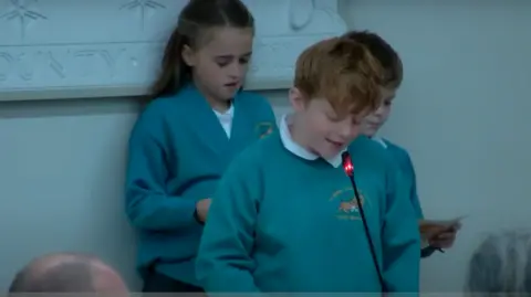 Hampshire County Council Two boys and a girl standing at a council meeting. They are wearing green school uniform tops and one boy is speaking into a microphone