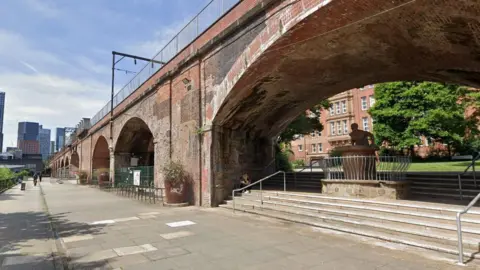 Google Red bricked archways loom over a street on a sunny day with tower blocks seen on the skyline in the distance. 