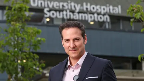 Cancer Research UK Dr Hugo stands in front of the University Southampton Hospital, which is in the background. He looks at the camera with his eyes narrowed. He is wearing a navy blue jacket and a pink shirt. There is a tree with green leaves between him and the hospital. 