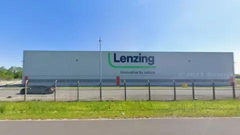 The exterior of Lenzing in Grimsby - a large, boxy, grey building with a large company logo on the front. It is behind a wire fence.