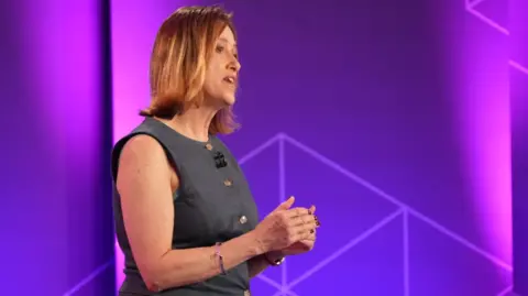 Jane Dodds standing in front of the purple backdrop of the TV set, holding her hands together and addressing the audience