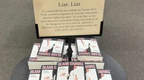 Brooke Jessop A pile of books on a table with a sign about the book in front of the books. You can see a large floor and the table is dark and round. 