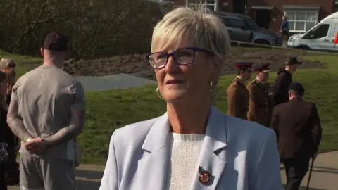 A woman with short grey hair and wearing glasses, a white jumper and pale lilac jacket with a military pin badge is talking to a reporter as they stand by the park.
