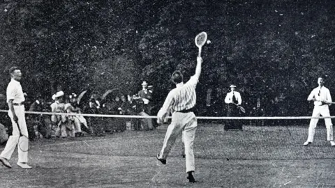 Getty Images A black and white image of a19th Century lawn tennis match. Three men and a woman hold wooden rackets, with a seated crowd sat by the sidelines.