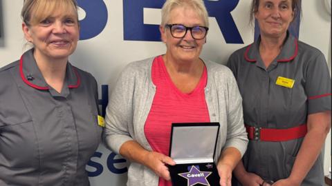 Three women stand closely together, smiling at the camera. The woman in the center, wearing glasses, a red top, and a light gray cardigan, holds an open display case containing a star-shaped award with the word 'Carer' on it. The two women on either side, dressed in matching gray uniforms with red trim and yellow name tags, stand proudly beside her.