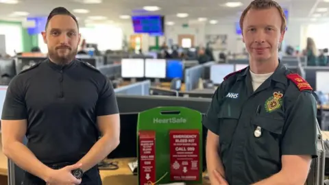 A police officer dressed in a black T-shirt with police epaulettes on the shoulder stands beside an ambulance worker dressed in his dark green uniform, with a red bleed kit between them. It has writing with instructions on it but it is not clear to read in this photo, beyond advice to call 999.