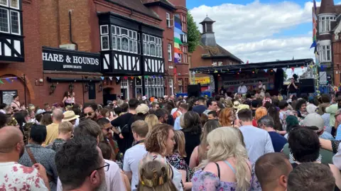 Picture shows hundreds of people on a street watching a stage at the far end where three musicans play. On the right hand side is the Hare and Hounds pub and its famous red brick and black and white facade. There is colourful bunting across the street and rainbow flags flying from buildings