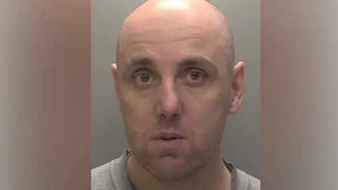 Humberside Police A police head shot of a bald man wearing a grey t-shirt. He is stood in front of a grey wall.
