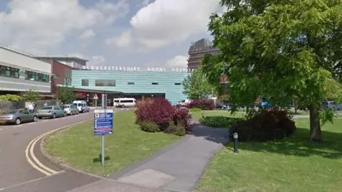 Google Image of the hospital entrance. The words Gloucestershire Royal Hospital appear in white above the turquoise tiles at the front ofthe building. a blue directional sign is in the foreground.